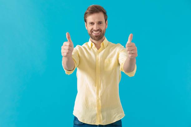 Positive casual man giving thumbs up and smiling, standing on blue studio background