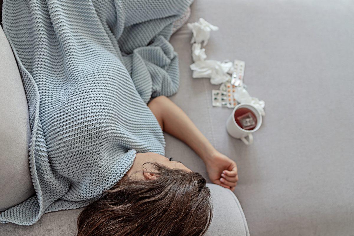 Sick Woman Sleeps Covered With a Blanket Lying in Bed With High Fever and a Flu, Resting at Living Room. She Is Exhausted and Suffering From Flu With Cup of Tea, Medicaments and Tissues